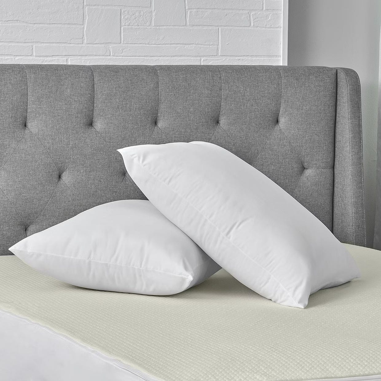 Two white pillows on a bed with a grey tufted headboard and a white brick wall in the background.