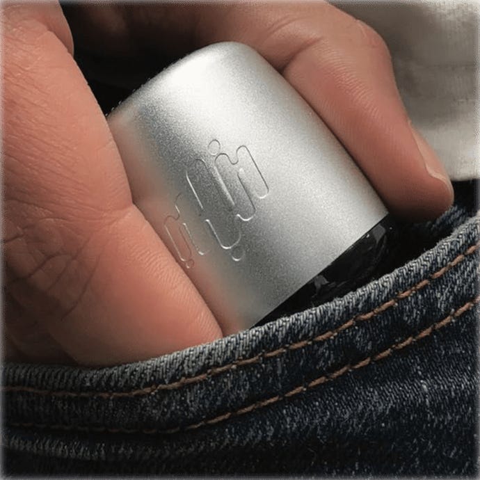 A silver cylindrical device with an embossed logo, being slipped into a denim pocket by a hand.