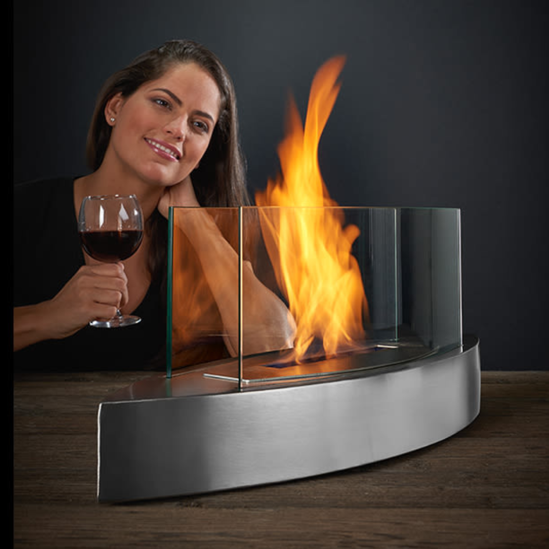 A modern tabletop fireplace with a sleek, curved stainless steel base and protective glass panels, featuring a bright, flame at its center.