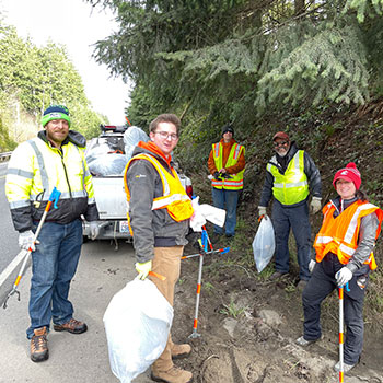 A group of Peninsula Subaru team members and Subie Adventure Club enthusiasts wearing safety vests and standing on the side of the highway with trash bags and trash pickers.