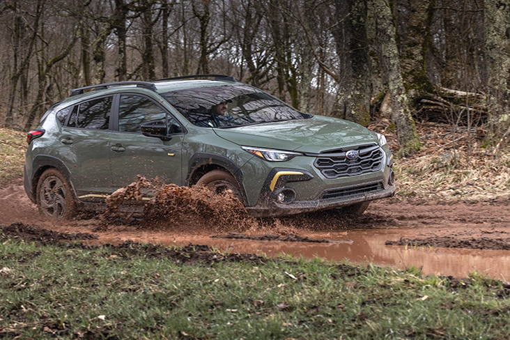 A 2024 Subaru Crosstrek Sport in Alpine Green is driving through a wet, muddy road in a forested area, splashing up water.