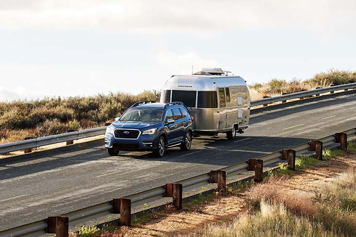An Ascent is traveling on a two-lane road with tall grasses beyond railings on both sides and is towing an Airstream trailer. Clouds can be seen in the distance with blue sky peeking through in patches.