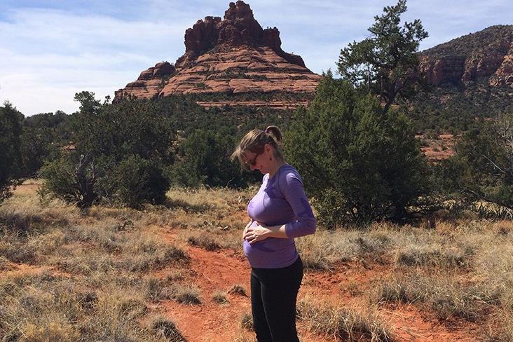 Catie Joyce-Bulay, seen from a side view, has her hands on her pregnant belly and is smiling. Behind her, Bell Rock formation reaches toward the sky on a sunny day in Sedona, Arizona.