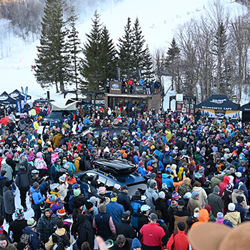 A huge crowd is gathered at Killington Mountain Resort in Vermont for Subaru WinterFest. Beyond the crowd, evergreen trees and a ski slope are visible.