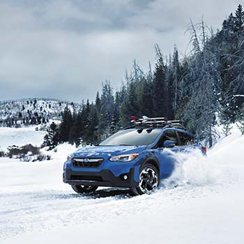 A 2022 Subaru Crosstrek in Horizon Blue Pearl is kicking up snow as it drives through an open area with evergreen trees in the background.