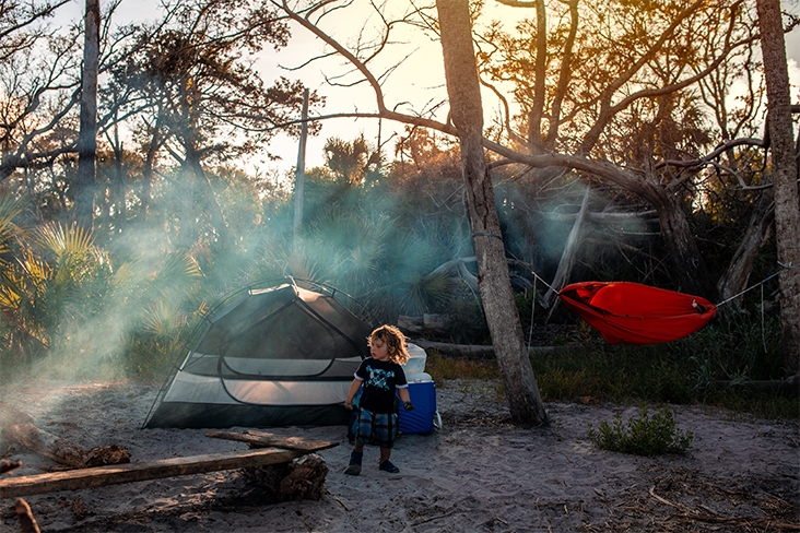 Jacobson’s child, who is standing in the sand, looking toward a smoldering campfire on the right. A tent and cooler sit behind the child, and a hammock is strung up between two trees beside the tent.