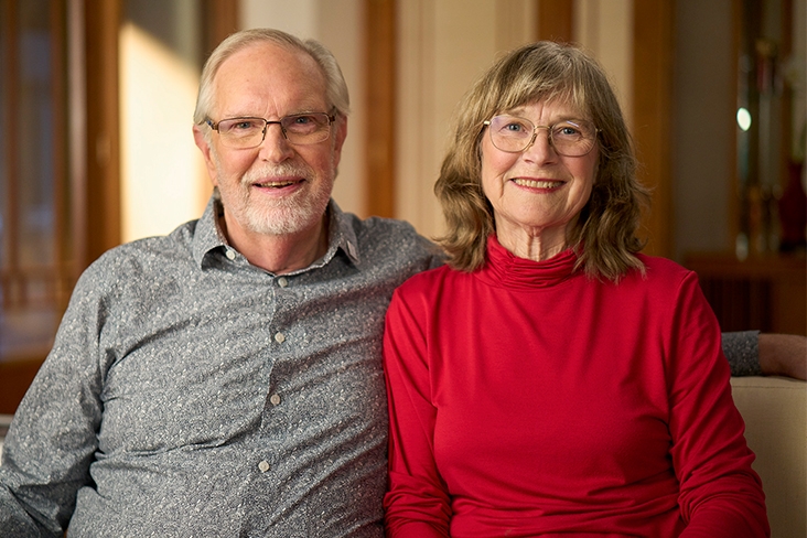 Don Nelms and his wife, Millie, are seated on a couch, smiling. Millie is wearing a red turtleneck while Don is wearing a button-down, long-sleeved shirt.