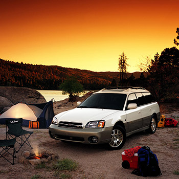 The Legacy Outback, which was a trim level of the Legacy Wagon. The vehicle is two-tone white and black and is located in a wilderness setting near water; two foldable chairs and a small fire are in front of it.