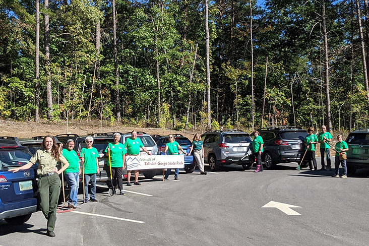 Several members of Friends of Tallulah Gorge State Park are standing in a parking lot next to their Subaru vehicles. Two members are holding a sign that reads Friends of Tallulah Gorge State Park.