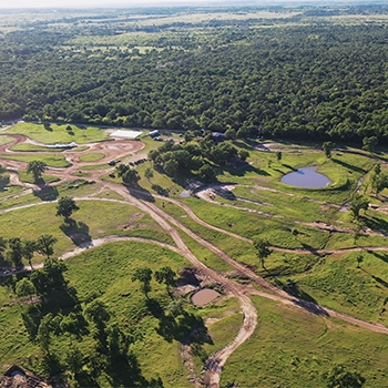 An aerial view of the Rally Ranch at Rally Ready. On the course, roads crisscross through greenery, a few patches of trees and a small pond. Behind the course is a thick line of deciduous trees, and they are fully leafed out.