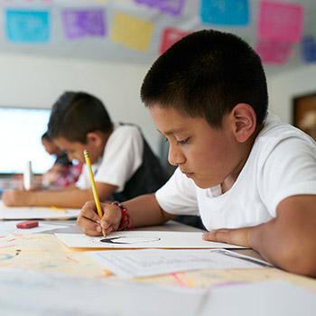 A young student is seated at a table in a classroom. He is writing with a pencil. The students next to him are also deep in thought, writing.