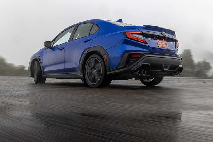 A wet asphalt road with a 2022 Subaru WRX driving past with the rear end of the vehicle showing