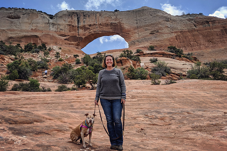 Stacy Dubuc and Ginger at Arches National Park in Utah. They are standing on a sandstone platform, and there is a massive natural sandstone arch in the background behind them.