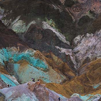 A shot taken from a distance of Artists Palette in Death Valley California. It reveals a colorful, rocky landscape, with purple and blue-green formations and people walking on top of them.