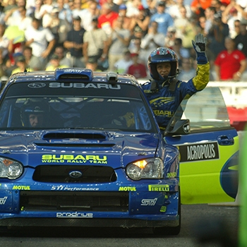 Petter Solberg is driving a World Rally Blue-painted Subaru vehicle at the Acropolis rally; he’s leaning out of the window and waving at the crowd with one hand while driving with the other. Chris Atkinson is next to him on the left.