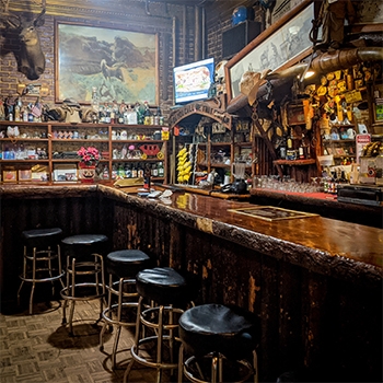 A bar in The Historic Virginian Hotel. The bar top appears to be crafted from long logs, and there are rustic-looking barstools underneath it. Western images are high on the wall and underneath the pictures are shelves filled with objects.