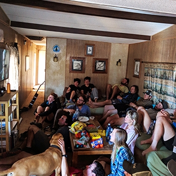 An older photo showing the living room before the remodel, with paneling and brown beams on the ceiling. The room is packed with about 15 guests watching TV.