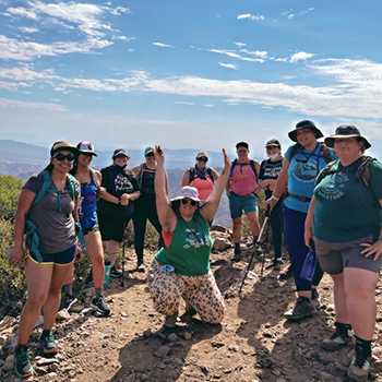 A group of happy Unlikely Hikers stand around Jenny Bruso as she crouches with arms raised along the Pacific Crest Trail in Cleveland National Forest just outside San Diego.