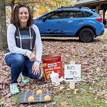 Lindsay Forney is in the outdoors kneeling next to some donations for pets; leaves are covering the ground. Her 2016 Subaru Crosstrek Limited is in the background.
