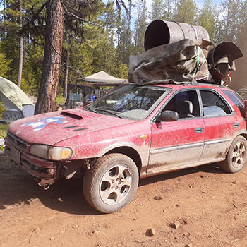A dirt-covered Subaru Impreza Outback Sport carrying scrap metal and other refuse on the roof is parked on a dirt road. Evergreen trees are close to the road and can be seen in the distance. 