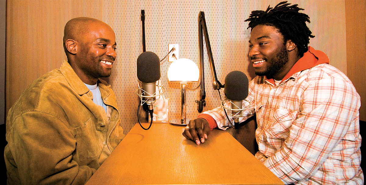 Two men are seated across from each other at a small table. They both have a large microphone hooked up in front of them, and they are relaxed and smiling.