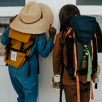 Two children are standing close to one another, and the view is from behind them. One is wearing a large straw hat and a backpack, and the other child is wearing a larger backpack and is holding a skateboard.