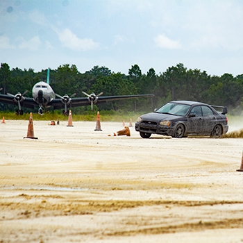 A car is racing on a dirt course around orange cones. In the distance, an airplane is grounded in a grassy field. 
