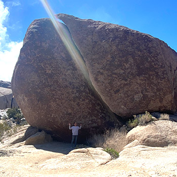 Nikko is standing underneath the overhang of an immense rock that is split vertically on Split Rock Trail in Joshua Tree National Park in the state of California.