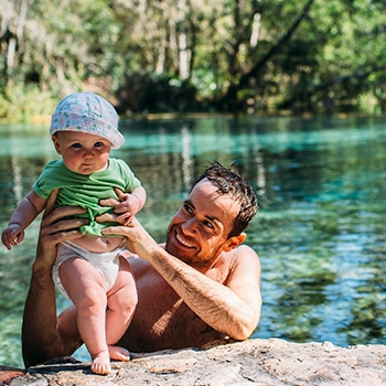 Joel is in the water of the springs, and we see him from the shoulders up, holding baby Land on the shore and smiling.