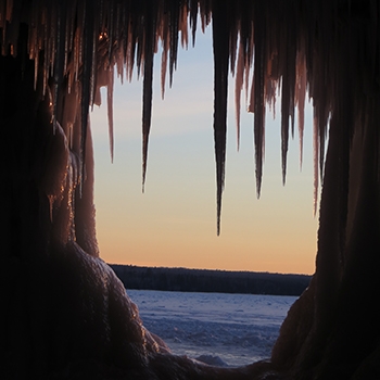 An ice cave entrance viewed from inside the cave. Sharp-looking icicles hang down from the entranceway.