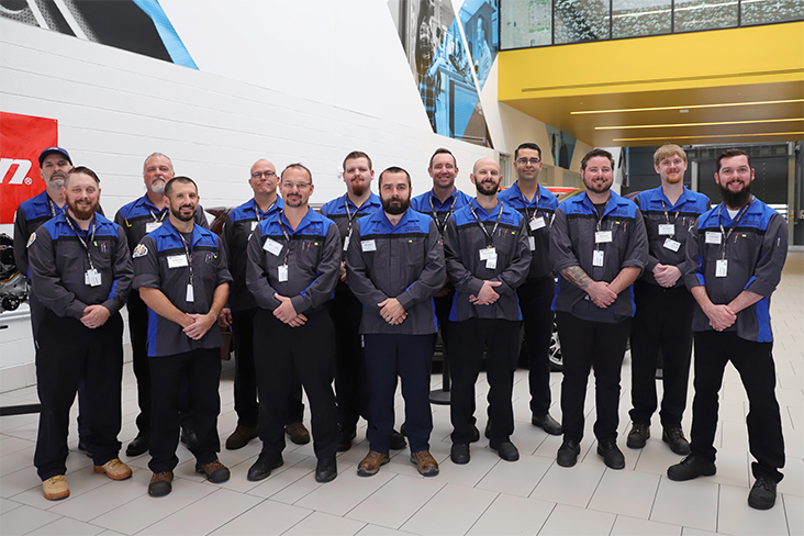 Fourteen men are standing in a group in a spacious area at Subaru headquarters in Camden, New Jersey, for the Subaru American National Technician Competition. They are wearing nametags on their shirts and automotive technician clothing for the competition.