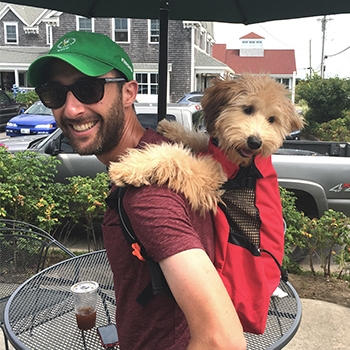Dan Saacks is wearing a green cap and red T-shirt. He's got Olive, the whoodle, strapped into a K9 Sport Sack Air 2 on his back. Olive has her paws resting on his shoulders and looks relaxed.