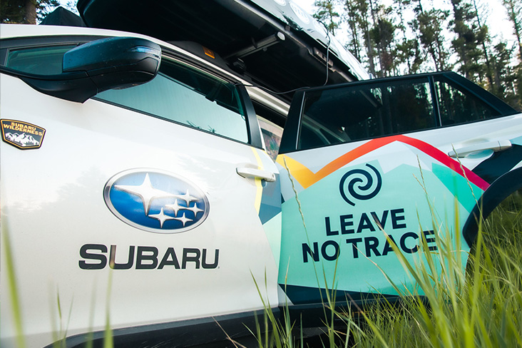 Close-up of the front end of a white Subaru Outback Wilderness vehicle. The front door says Subaru and has the Subaru logo. The back door says Leave No Trace and has the Leave No Trace logo. 