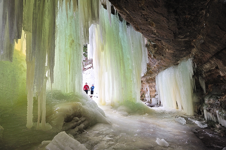 Two hikers entering the mouth of Eben Ice Caves. They are standing in between floor-to-ceiling waterfall-like ice that glows a blue-green color.