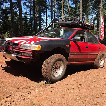 A modified Subaru with rally tires on a dirt road in a forest. The front panel is painted black while the back panel is red.