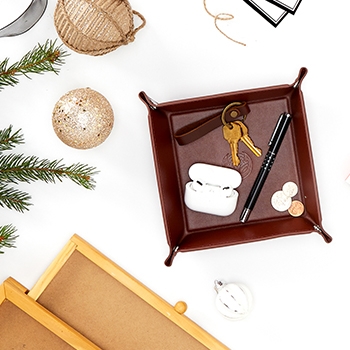 The mahogany-colored valet tray with keys, a pen and coins inside of it. Decorative ornaments and pine needles placed near the tray provide a festive look.