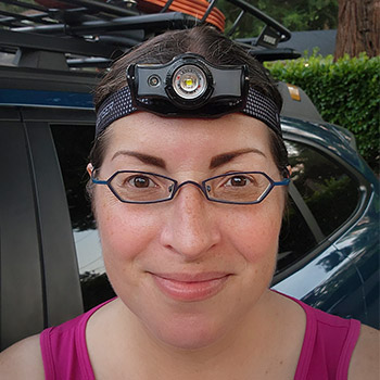 Writer Mercedes Lilienthal, a brunette woman with brown eyes and glasses, smiles while wearing a Ledlenser headlamp across her forehead.