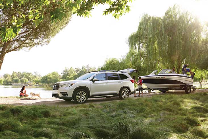 A Subaru Ascent Limited in Crystal White Pearl with a boat hitched on the back is parked on a gravel road by a lake. The rear gate is up and under it is a young woman who is securing a life jacket on an adolescent male. Tall grass is in the foreground, a young woman is seated near the water with a small white dog, and there are mature deciduous trees in the distance circling the water.