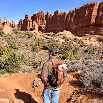 The writer’s husband, Jason, hikes in Arches National Park in Utah. He is wearing a backpack with the plush jack rabbit sitting inside it with its head appearing to look out.