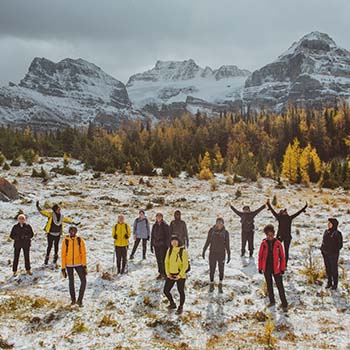 A group of hikers in a clearing at Larch Valley Trail in Alberta, Canada, with evergreen trees and mountains in the background. Some are waving their arms in the air, and theyʼre all wearing jackets for cooler weather.
