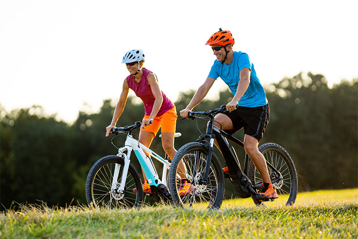 A man and a woman are e-biking side by side on a grassy path. Blurred trees can be seen in the background. They are wearing helmets and summer clothing and are smiling.