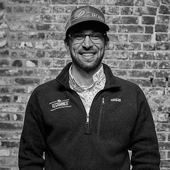 ReGrained co-founder Dan Kurzrock is wearing a cap and pullover jacket and standing in front of a brick wall, smiling.