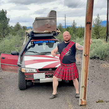 Brent Burbank, a Gambler 500 competitor, is wearing a kilt and standing in front of the modified Subaru that he used in the rally.