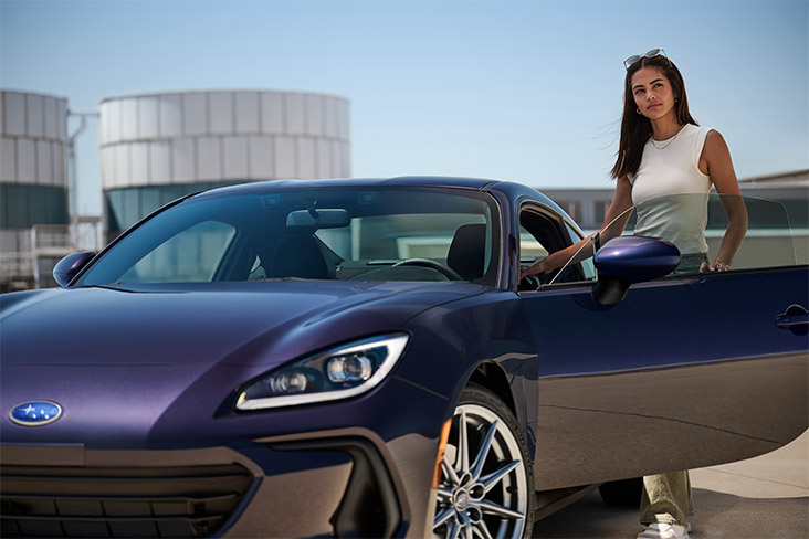 The 2025 Subaru BRZ Series.Purple is parked on cement with the driver's side door open and an attractive model standing behind the door with sunglasses on her head.