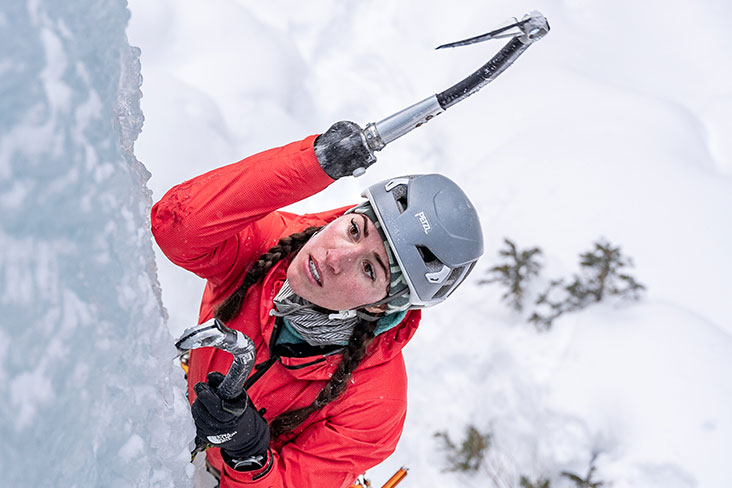 Closeup of Kimber Cross as she climbs up a wall of ice using her prosthetic device with a look of concentration on her face. She is wearing her hair in two braids and has on a gray helmet and a red winter jacket.
