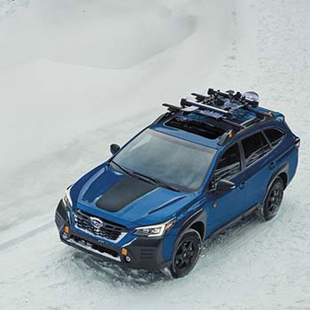 Aerial view of a Geyser Blue Subaru Outback Wilderness driving on a snow-covered road. There is a Thule Ski and Snowboard Carrier on the rooftop.