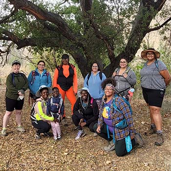A group of members from the Los Angeles chapter are posing underneath a tree during their first hike in 2021. There are six people standing and four more kneeling in front of them. They are smiling and wearing wide-brimmed hats or caps.