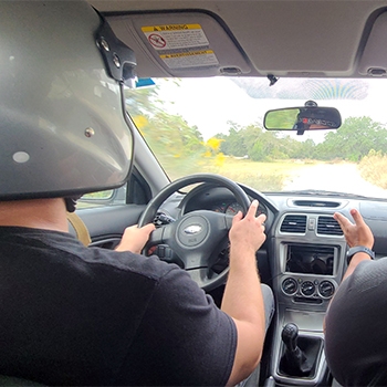 A shot of a driver behind the wheel of a car at RallyPro. He’s wearing a gray helmet, and his hands are both on the steering wheel. There is also a person in the passenger seat, but from the camera’s angle, only his shoulder and arm are visible.