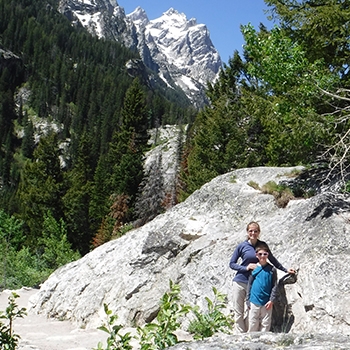 Casey and Caden at Grand Teton National Park. Casey is standing behind Caden, and she has her hand resting lightly on his shoulder. They are standing in front of a large boulder, which is much taller and wider than they are, and ragged mountaintops, evergreen trees and a blue sky are in the distance.