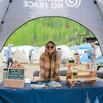 A female is standing behind a Leave No Trace table in a tent. The table has handouts and there is a small chalkboard that says, Are you camping during the festival? There are many tents set up behind her in the background.
